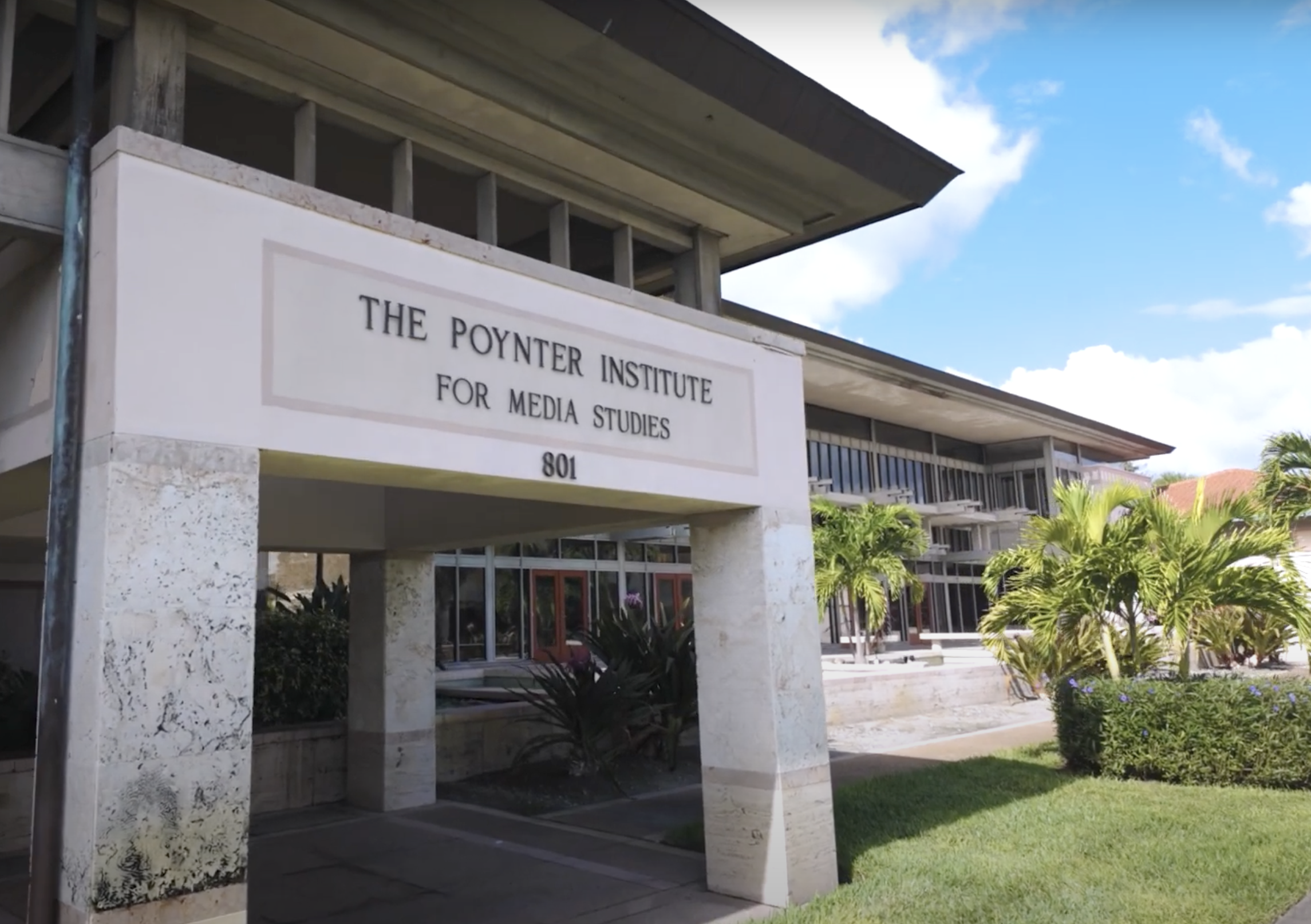The Poynter Institute, beige building surrounded by green grass and palm trees on a sunny, slightly overcast day.